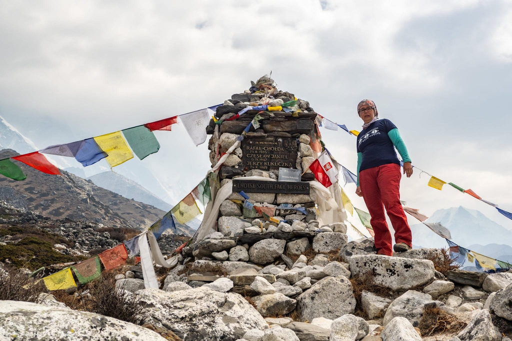 Kobieta stoi obok kamiennego pomnika w g&oacute;rach, otoczonego kolorowymi flagami modlitewnymi.